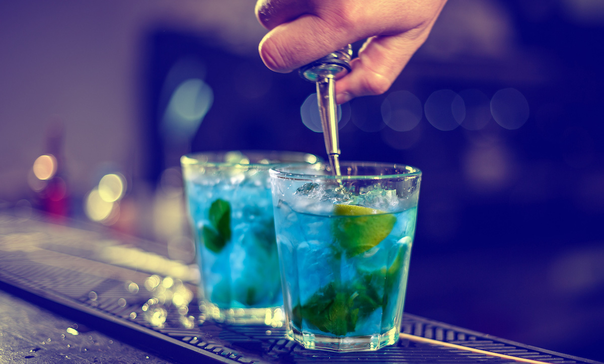 Bartender is pouring blue syrup into the glass with ice cubes on the bar counter